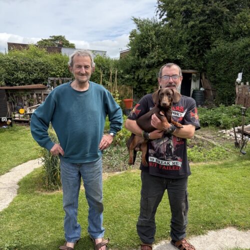 Two men stood in their garden with a daschund dog